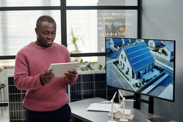 Leinwandbilder - Young serious businessman looking at tablet screen against his workplace with interactive board and models of windmills and house