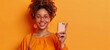 A lively and smiling young woman with afro hair and glasses, excitedly pointing her finger up and holding a smartphone in the same hand, on an energetic yellow background. Copy space.