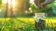 Saving Money in a Jar on Sunny Grass. A hand placing a coin into a glass jar full of coins on lush green grass, bathed in warm sunlight, symbolizing savings and financial growth.