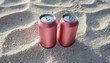 2 pink aluminum can with condensation drops on clear white sand at beach. Beer or soda drink package.
