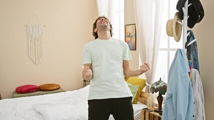 Wall Mural - Excited young caucasian man in pyjamas jumps in joy, screams with winner's gesture celebrating success in bedroom