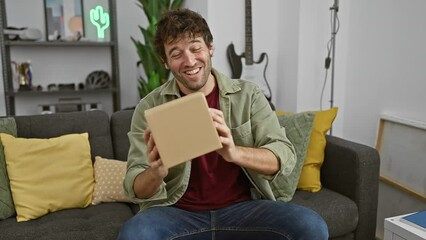 Canvas Print - A cheerful man holds a package in a cozy living room, expressing excitement and satisfaction with his delivery.