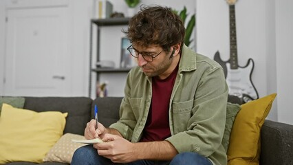 Poster - A young hispanic man with a beard writes notes at home, showcasing casual indoor living with a thoughtful expression.