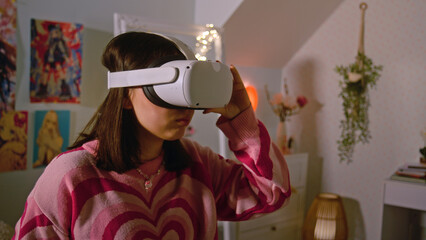 Teenage girl wearing AR headset plays virtual reality online video games in her cozy bedroom. Girl spending leisure time and having fun at home. Concept of modern devices, entertainment and lifestyle.