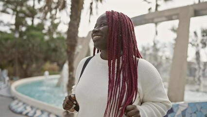 Sticker - A smiling african woman with braids in a sweater posing by a fountain in a park