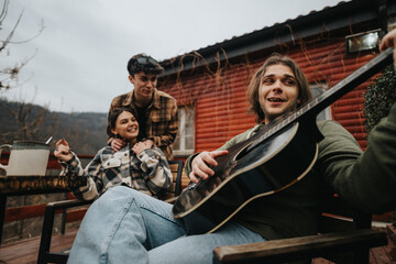 Wall Mural - Group of young adults smiling and bonding over a guitar play along on a rustic wooden porch, exuding a sense of leisure and friendship.