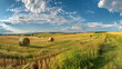 Expansive view of lush green farmland with rolls of hay bales under a clear blue sky with fluffy clouds stretching across the horizon