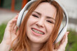 Portrait of a young red-haired woman with braces on her teeth listening to music on headphones outdoors. 