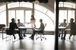 © fizkes - Small diverse business team discussing project at meeting table in office work space, sitting and standing at large window and daylight. Young manager woman presenting ideas. Full length wide shot