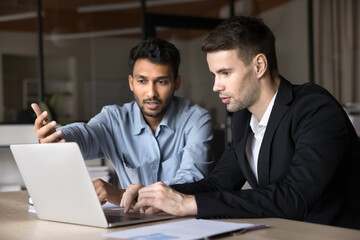 Two diverse business men talking at laptop, discussing online job application, Internet project, startup, looking, pointing at computer screen. Indian manager, presenting web content to customer
