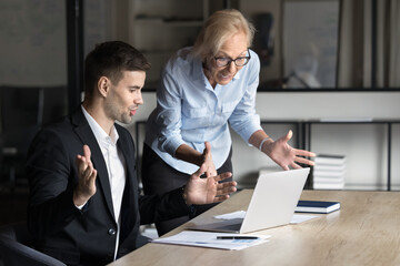 Canvas Print - Two excited worried younger and elder business colleagues using laptop at workplace, staring at screen in surprise, speaking getting email, notice, message with unexpected concerning news