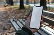 A hand holding a smartphone with a white mockup screen on a park bench