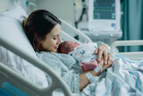 Fototapeta Londyn - Cheerful new mother carrying and smiling to her newborn baby while resting on the hospital bed after giving birth