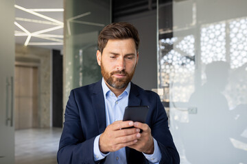 close-up photo of a young serious male businessman who is in a modern office and uses a mobile phone