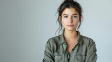 A Portrait Of A Young Woman With Natural Makeup, Wearing A Casual Olive Green Shirt, Looking Directly At The Camera With A Neutral Expression Against A Plain Grey Background.