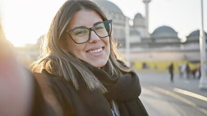 Poster - A joyful woman with glasses smiles and poses playfully in the historic streets of istanbul during a bright sunset hour.