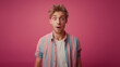 A young blonde man in striped shirt surprised and shocked looking at camera, doing silly face, on a pink background