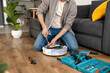 man repairs robot vacuum cleaner and tightens bolt with tools sitting on wooden floor in blue jeans and gray shirt. Scheduled service and cleaning in a room with a gray sofa and a green vase