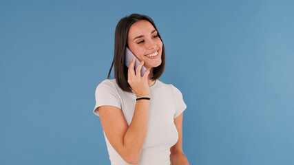 Wall Mural - Laughing beautiful young brunette woman dressed in white top talking on smartphone isolated on blue background in studio