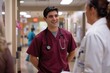 © EarthWalker - Male nurse practitioner wearing maroon scrubs and white coat talking to colleagues at work in hospital. AI Generated