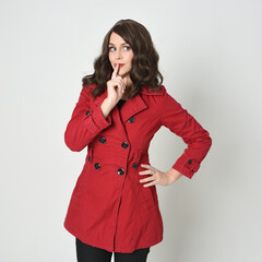 Wall Mural - close up portrait of beautiful brunette woman model, wearing red trench coat jacket.
isolated on white studio. finger half to mouth in secretive shushing gesture.