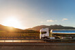 Truck with a food tanker driving next to a field of vineyards with the sun rising between the mountains, side view.