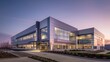 © kittikunfoto - Modern office building at twilight with illuminated interior windows, landscaped surroundings, and an empty parking lot.