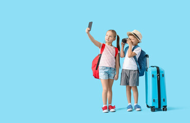 Wall Mural - Cute little children with backpacks, suitcase and camera taking selfie on blue background. Travel concept