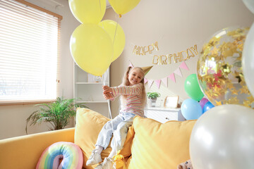 Canvas Print - Cute little girl sitting on sofa in party hat with balloons, toys and decorations for birthday at home