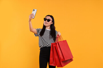 Wall Mural - Smiling woman with shopping bags taking selfie on yellow background