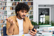 © Liubomir - A young adult man looks puzzled while reading something on his smartphone, surrounded by a stack of books, possibly studying.