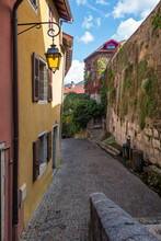 A Narrow Street With Old Houses Along The Wall Of A Medieval Castle, Overgrown With Climbing Plants. There Is A Lantern And Dark Paving Stones. Annecy. France.