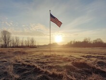 A Large American Flag Is Flying In A Field. The Sky Is Clear And The Sun Is Setting, Casting A Warm Glow Over The Grassy Field. The Flagpole Is Tall And The Flag Is Large