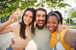 © CarlosBarquero - Selfie three excited multiracial joyful young friends embracing outdoors. Women and man having fun looking at smiling camera and making peace symbol hands enjoying summer vacation. Gen z in sunny park