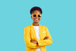 Fashion model wearing stylish suit and sunglasses. Happy smiling attractive young African American woman in white top, yellow jacket and glasses posing with arms folded isolated on blue background