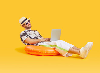 Business entrepreneur or remote employee working on computer on summer holiday trip vacation. Happy funny tourist man typing on laptop PC, lying on comfortable orange ring yellow studio background