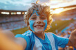 Exuberant boy taking selfie with blue face paint at a football match, feeling excitement and joy