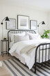 A black metal bed frame in the middle of an all-white bedroom with three frames on the wall above it, a striped grey rug, and two lamps on side tables, a modern style bedroom.
