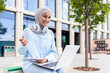 Focused young student wearing a hijab with headphones studying on her laptop and taking notes outside on campus.