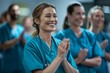 A cheerful female healthcare professional in blue scrubs clapping hands with her colleagues in the background.