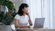 © Wasana - Asian woman sitting contemplating holding a coffee cup while looking away from her laptop. Looking out of the glass window In the relaxing corner of the cafe with green trees