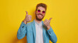 A happy young man with a beard on a yellow background smiles and shows thumbs up