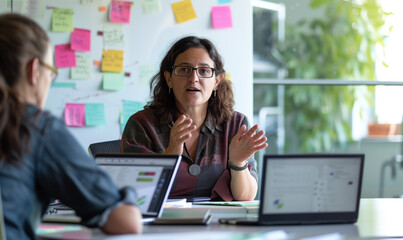 Wall Mural - Dynamic Business Discussion. An animated conversation unfolds as a woman in her late thirties, with glasses, discusses project details in a vibrant office setting.