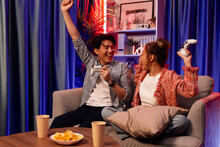 Couple Gamer Raising Fist Up Holding Joysticks Be Winner Together Complete Level Sitting Sofa Playing Video Game With Front Snack And Drinks At Neon Light Color Living Room At Modern Home. Infobahn.