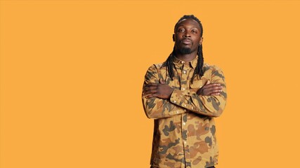 Wall Mural - Portrait of guy with dreads smiling in studio set, wearing trendy camo clothes and feeling joyful on camera. African american adult with cool hair posing over orange background, natural style.