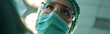 © eggeeggjiew - Low angle view of a focused female surgeon looking up, framed by the glare of surgical lights during an operation