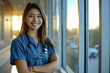 Radiant young healthcare professional in blue scrubs, arms crossed and smiling in a sunlit hospital hallway