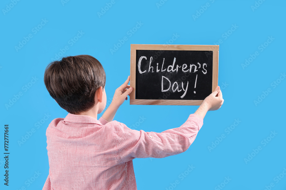 Little boy holding chalkboard with text CHILDREN'S DAY on blue background, back view