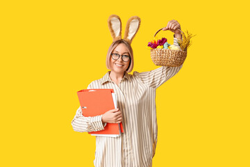 Wall Mural - Happy adult female secretary in bunny ears with documents folder and basket for Easter on yellow background