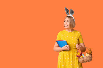 Wall Mural - Happy adult female manager in bunny ears with notebooks and basket for Easter on orange background